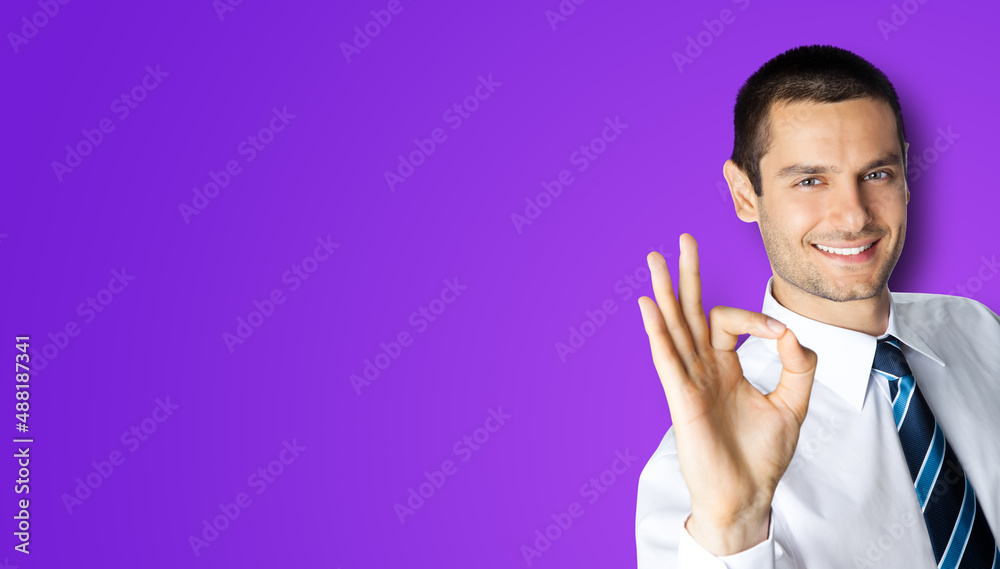 Portrait of smiling businessman in white shirt and tie, showing thumb up like hand sign gesture, isolated on violet purple background. Happy confident man gesturing. Business success concept.