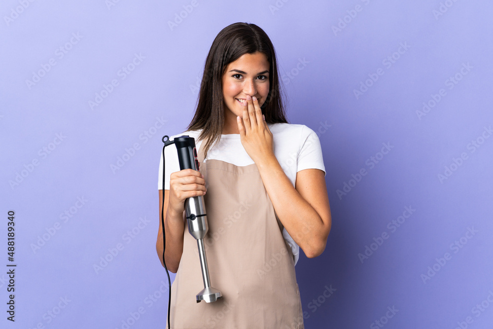 Brazilian woman using hand blender isolated on purple background happy and smiling covering mouth with hand Brazilian woman using hand blender isolated on purple background happy and smiling covering mouth with hand