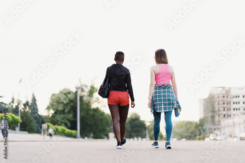 Friends back view in sportswear walking after a sport session in the city discussing . Multiethnic women having a fitness workout jogging.