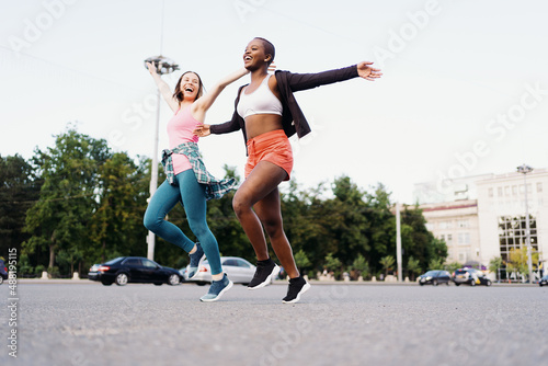 Cheerful smiling friends in sportswear running in the city dicussing. Multiethnic women having a fitness workout. photo