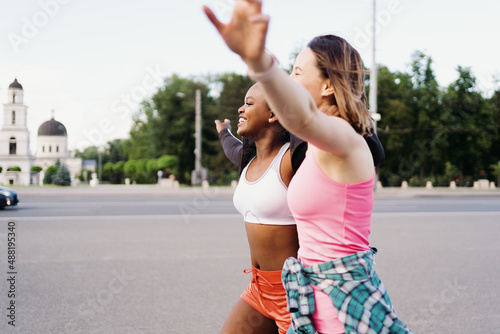 Cheerful smiling friends in sportswear running in the city dicussing. Multiethnic women having a fitness workout. photo