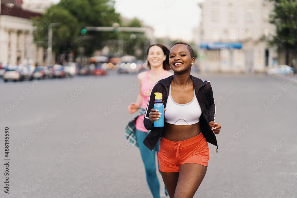 Cheerful smiling friends in sportswear running in the city dicussing. Multiethnic women having a fitness workout. Cheerful smiling friends in sportswear running in the city dicussing. Multiethnic women having a fitness workout.