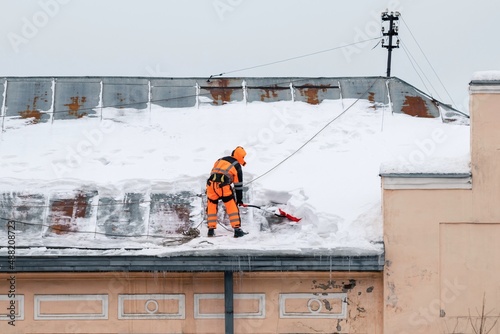 A working man in bright overalls with a safety belt with a shovel clears snow from the roof of an old building. Prevention of snow falling from the roof, industrial mountaineering