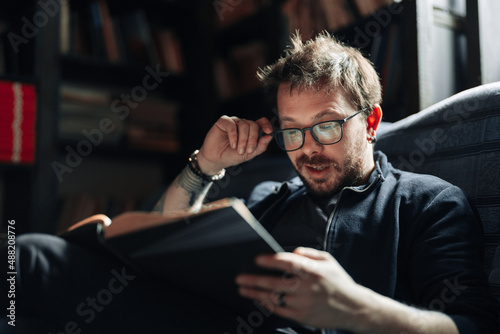 Adult student reading a book in the college library. Young positive male wearing glasses with bookshelves on background.