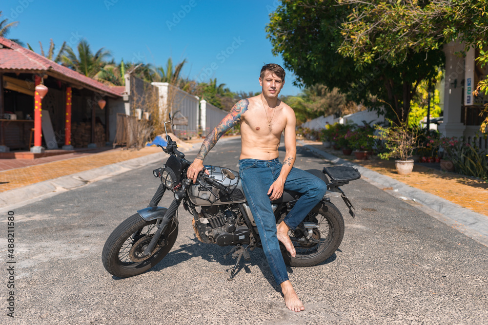 Fototapeta premium Young shirtless caucasian man with full-arm tattoo sitting on motorcycle
