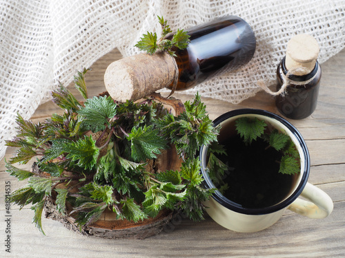 Quadro em tela Fresh young nettle leaves, herbal drink in a mug, juice, tincture in a bottles on a wooden background, top view