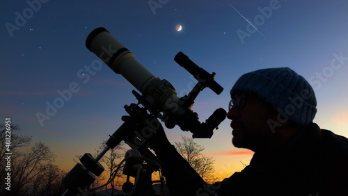 Silhouette of a man, telescope and countryside under evening skies.