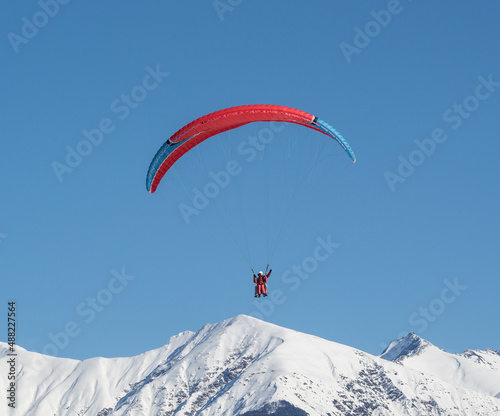 People in tandem paragliding over winter snowy mountains at clear blue sky background