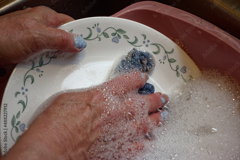 Fototapeta premium A woman's hand, with newly manicured nails, still does the dishes by hand in a washbasin in the Kitchen Sink.