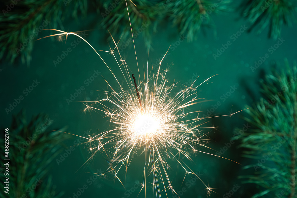 Obraz premium Burning sparkler with sparkles on a long shutter speed. Dark green background decorated with branches fir tree and pine