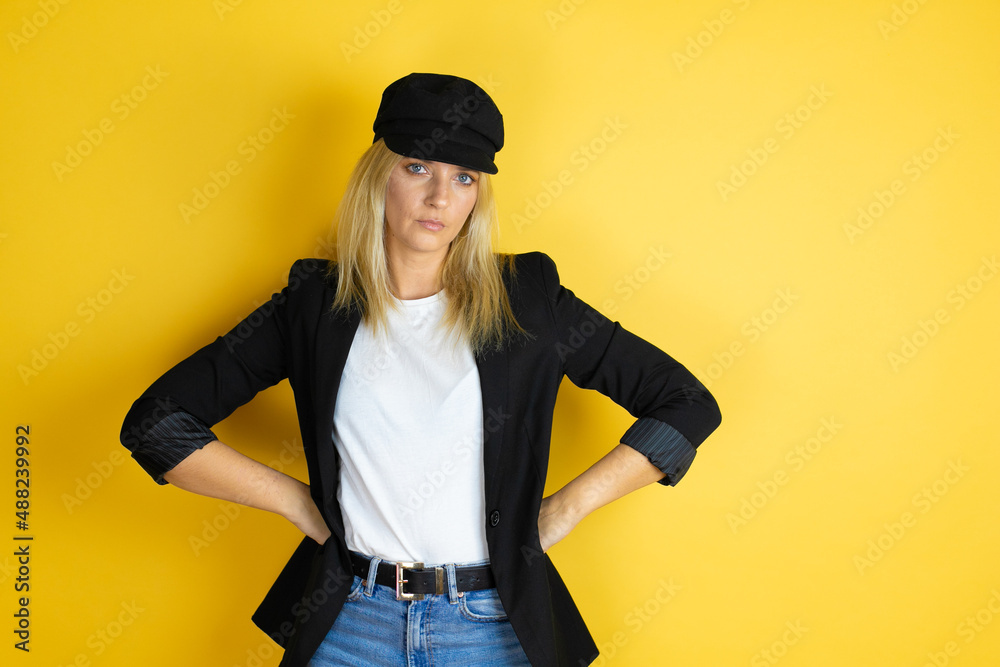 Beautiful woman wearing casual white t-shirt and a cap over isolated yellow background skeptic and nervous, disapproving expression on face with arms in waist Beautiful woman wearing casual white t-shirt and a cap over isolated yellow background skeptic and nervous, disapproving expression on face with arms in waist