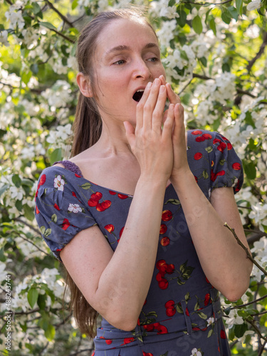Young woman sneezes in the park against the background of a flowering tree. Allergy to pollen concept.