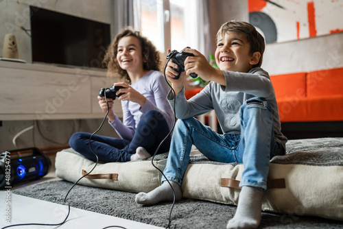 Two children small caucasian brother and sister happy children siblings boy and girl playing video game console using joystick or controller while sitting at home real people family leisure concept photo