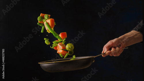 A frying pan in the chef's hand with pieces of red fish and vegetables on a dark background. Levitation. Sea food. Dishes from fish and seafood. Recipes, cookbook, food blog.