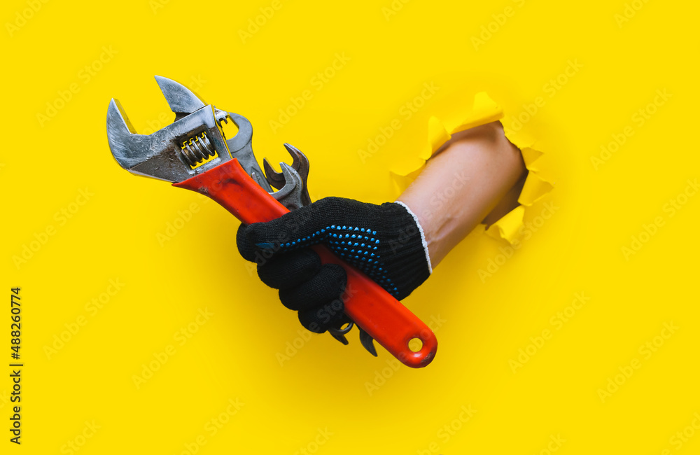 Naklejka premium The right hand, in a black knitted glove, holds several wrenches and an red adjustable wrench. Torn hole in yellow paper. The concept of a worker, a labor migrant, a master of his craft. Copy space.