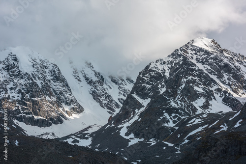 Dramatic landscape with sunlit snowy mountain range in low cloudy sky. Golden shine on white snow and black rocks in low clouds. Awesome view to snow mountains with gold sunlight in changeable weather