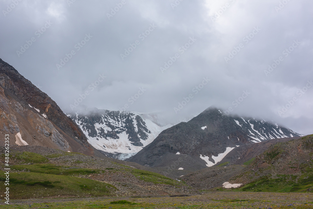 Gloomy landscape with snow mountain range in gray low clouds in overcast. Atmospheric mountain scenery with snow mountains among low clouds. Dramatic view to mountains under gray sky in rainy weather.