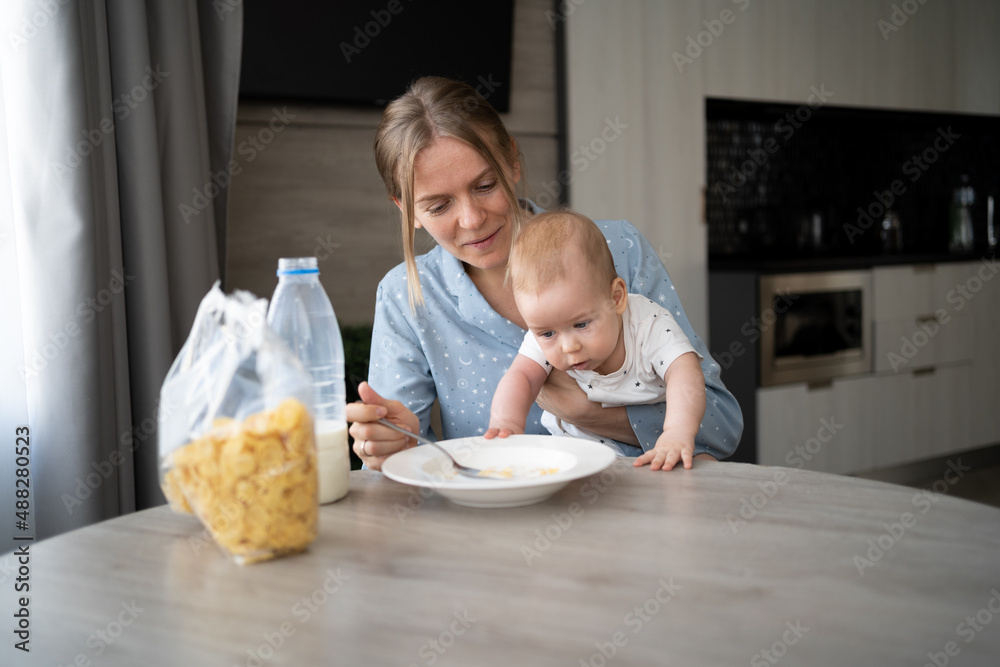 Family having breakfast in a white sunny kitchen. A young mother with a baby son eats corn flakes with milk, food for children. Parent with toddler and child Family having breakfast in a white sunny kitchen. A young mother with a baby son eats corn flakes with milk, food for children. Parent with toddler and child