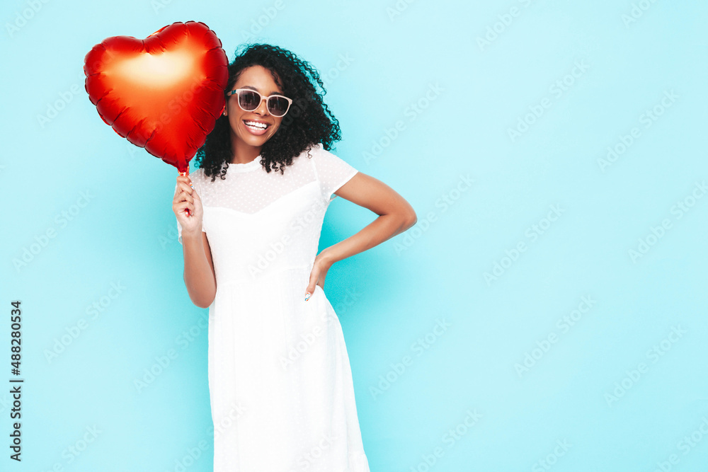 Beautiful black woman with afro curls hairstyle. Smiling model dressed in white summer dress. Sexy carefree female posing near blue wall in studio. Tanned and cheerful. Holding heart air balloon