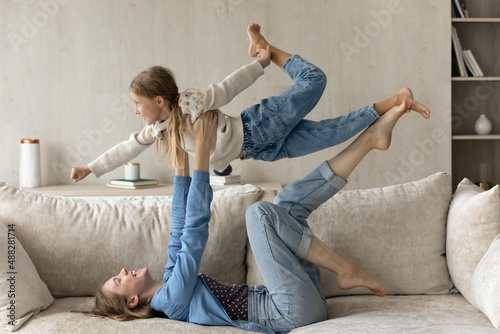 Joyful happy mom holding daughter kid up in air on hands and leg base, lying on back, resting on comfortable couch, playing active sportive games with child, doing acrowiga support