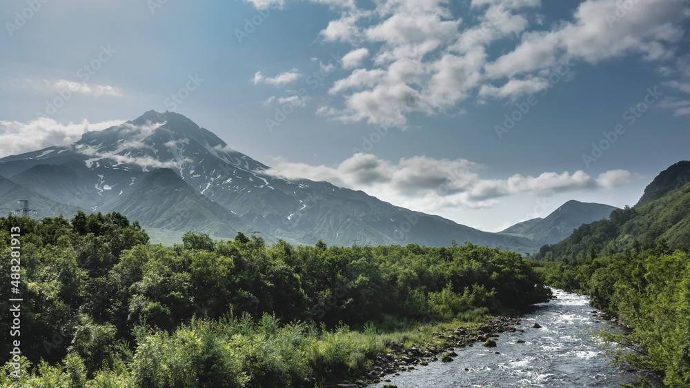 A mountain river boils and foams on a rocky bed. Lush green vegetation on the banks. A beautiful conical volcano on a background of blue sky and clouds. Kamchatka. Stratovolcano Vilyuchinsky