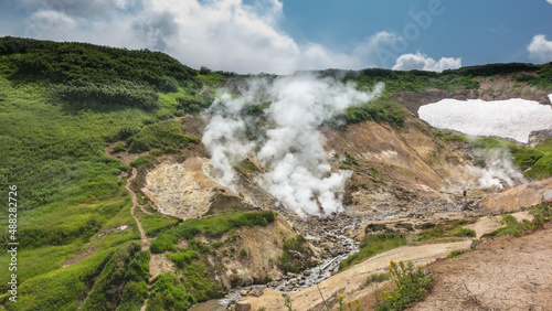 A stream from hot springs flows among the stones. Clouds of steam and smoke from fumaroles rise over the slopes of the mountains. Green vegetation and areas of snow around. Blue sky. Kamchatka.