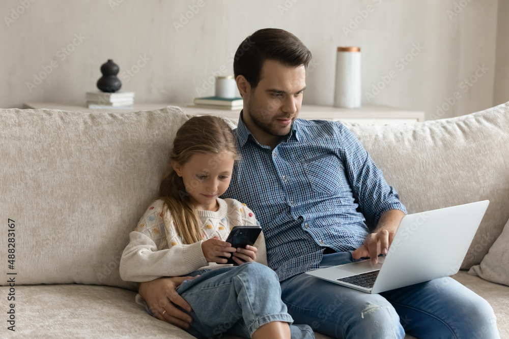 Focused digital addicted dad and gen Z daughter kid engaged in gadgets using. Father and girl resting on couch, browsing internet, social media on laptop, playing virtual video game on smartphone