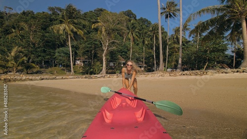 Young woman and sunglasses pulls pink plastic canoe with paddle to azure sea water from beach with palms. The beginning of the boat trip, boarding. Traveling to tropical countries