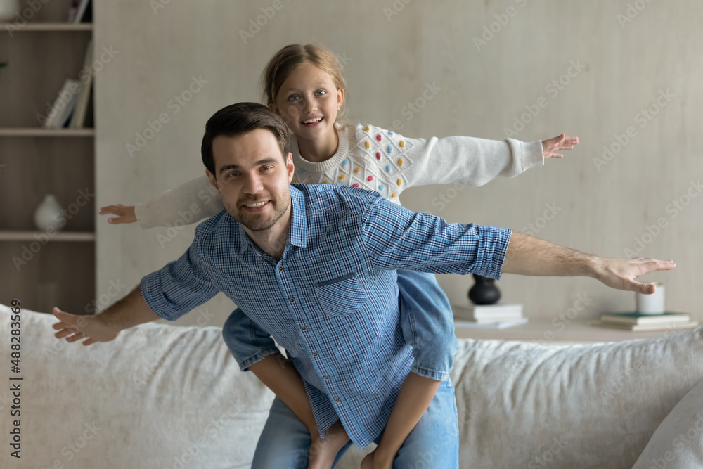 Happy millennial daddy and pretty daughter kid playing active games at home, having fun, imitating airplane with open flying arms, looking at camera, smiling. Dad piggybacking child. Family portrait