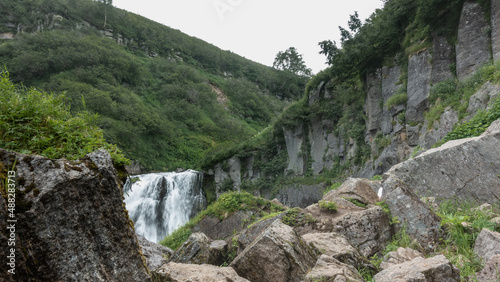 The streams of the waterfall collapse from the edge of the plateau into the gorge. There is green vegetation on the mountain slope. In the foreground are picturesque boulders. Kamchatka
