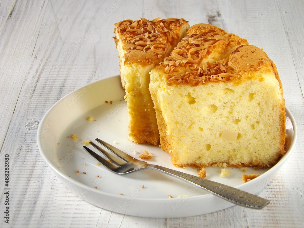 Sliced moist cheese chiffon sponge cake served in white plate on crochet place mat over the white wooden background. Copy space available. Sliced moist cheese chiffon sponge cake served in white plate on crochet place mat over the white wooden background. Copy space available.