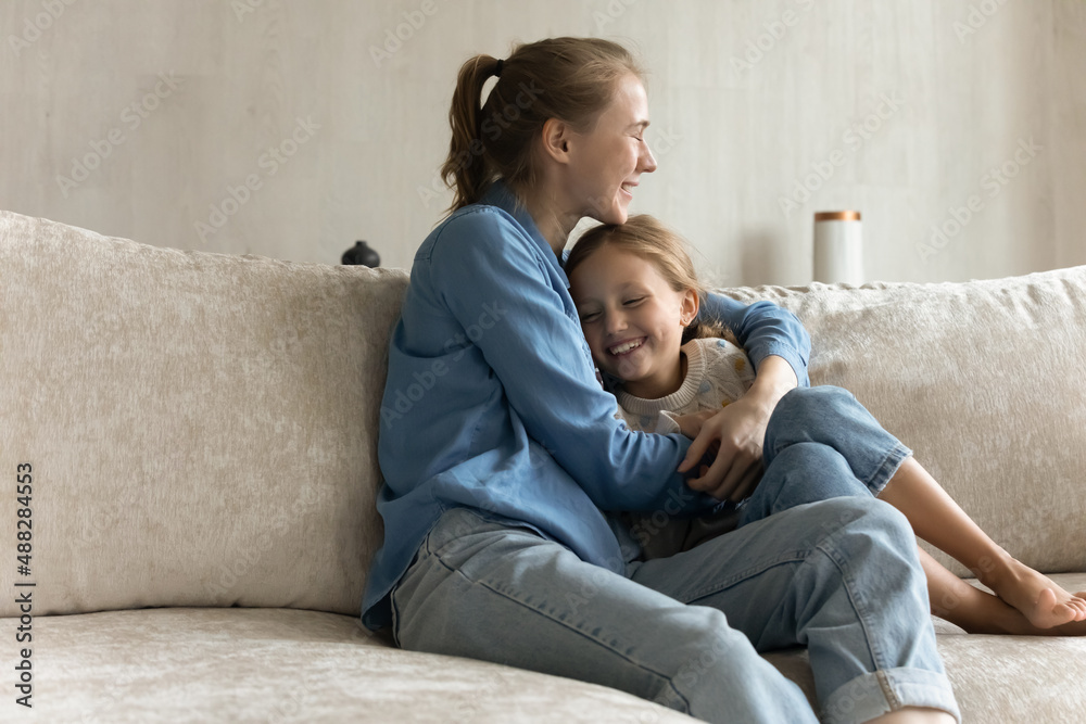 Happy cheerful mom hugging laughing daughter kid with love, tenderness, gratitude, sitting on comfortable couch at home, enjoying motherhood, closeness with child, family leisure time