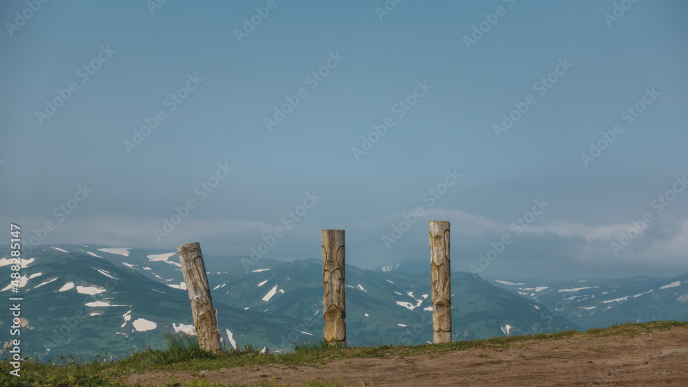 Old wooden totem poles stand on the hillside. Background - blue sky, mountain range. Copy space. Kamchatka. Vilyuchinsky Pass