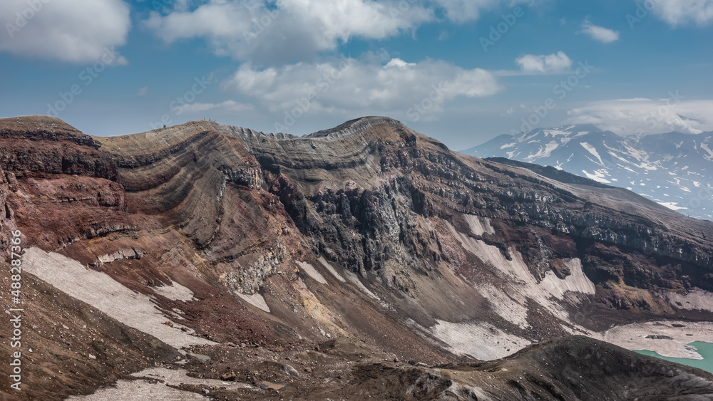 The edge of the volcano crater. Close-up. Layered structure of slopes, spots of melted snow, a section of acid lake. Tiny figures of people against the blue sky. Kamchatka.  Gorely Volcano