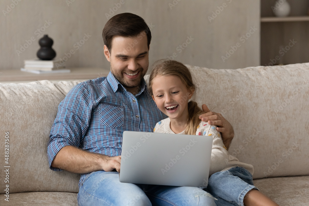 Happy daddy and cheerful daughter kid relaxing on couch at home together, using online learning app on laptop, watching online movie, series, TV channel, talking on video call, laughing