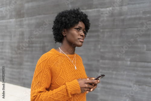 Tela Young afro woman holding a mobile phone while walking on the street