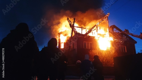 The roof of the house is on fire. The residential building burn, village. Firefighters put out a fire from the crane from above. The fire Department putting out the open flame. Silhouette of people. © ivandanru