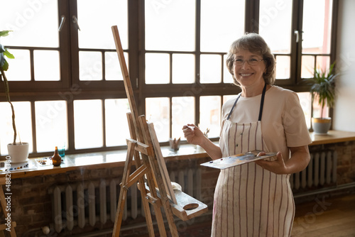 Portrait of smiling positive old senior retired woman in eyeglasses wearing painted apron, standing near wooden easel, feeling inspired drawing artwork on canvas in modern loft studio or living room.