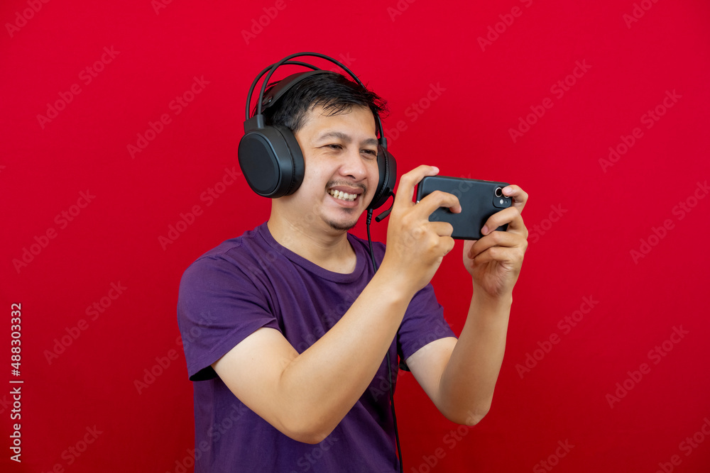 An asian man is playing a game on his smartphone and wearing a headset. An asian man is playing a game on his smartphone and wearing a headset.