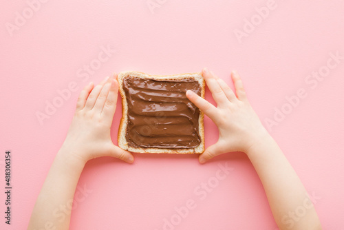 Baby girl hands touching fresh slice of white bread with brown chocolate cream on light pink table background. Pastel color. Closeup. Point of view shot. Sweet snack. Top down view.