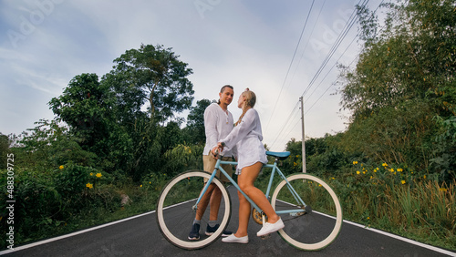 Biking road trip. Love couple on blue bike in white clothes on forest road. Just married woman and man kiss, hugs, stand on bicycle. Wedding, honeymoon. Cycling Cycle Fix. Asia Thailand ride tourism. photo