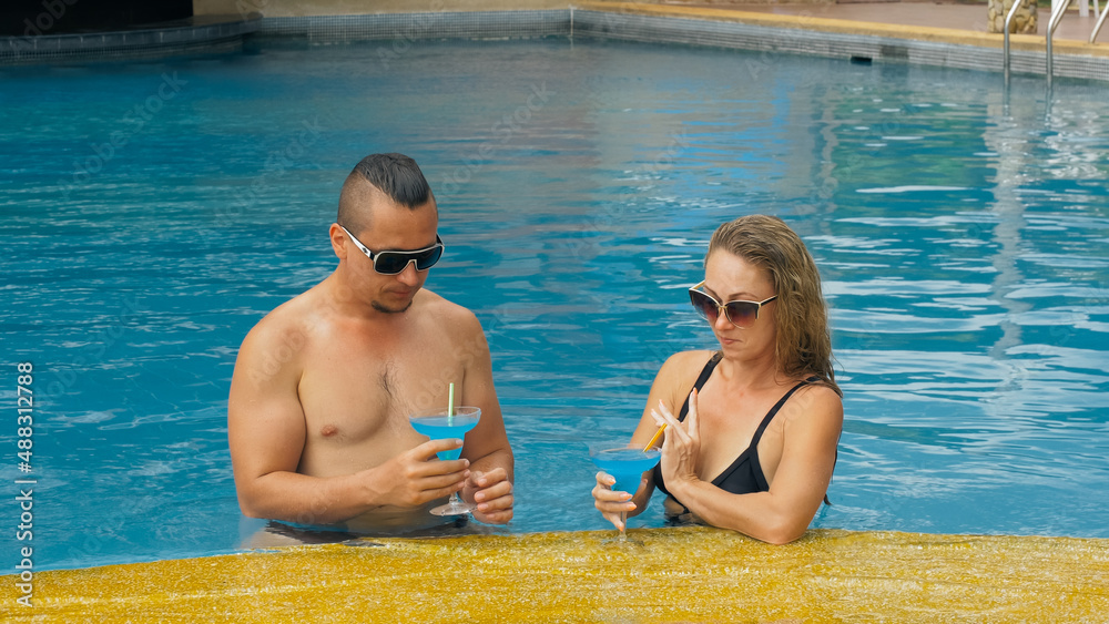 The loving couple hugs and kisses, drinking blue cocktail alcohol liquor in swimming pool at hotel outdoor. Portrait of caucasian man and woman. Creative hairstyles bodybuilder, swimsuit, sunglasses.