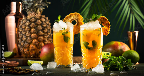 Alcoholic cocktail with vodka, pineapple juice, mango, liquor, ice. Long drink or summer mocktail. Tropical dark background with palm leaves and exotic fruits