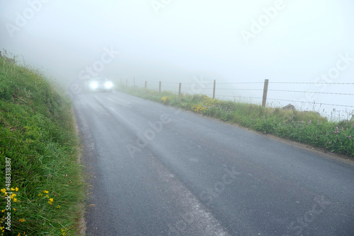 Car with headlights on on a small asphalt country road in a fog. Road safety and travel in dangerous weather conditions concept.