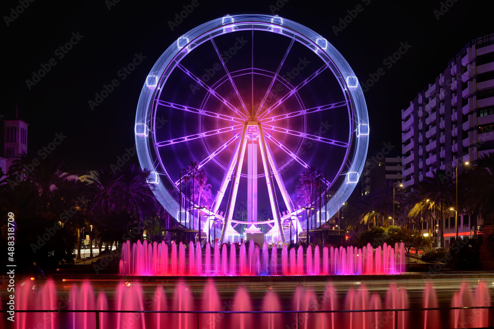Long exposure photo of a Ferris wheel ride in operation behind water fountain jets in the city, surrounded by palm trees and beautifully colored buildings on fair night. Fun. Entertainment.