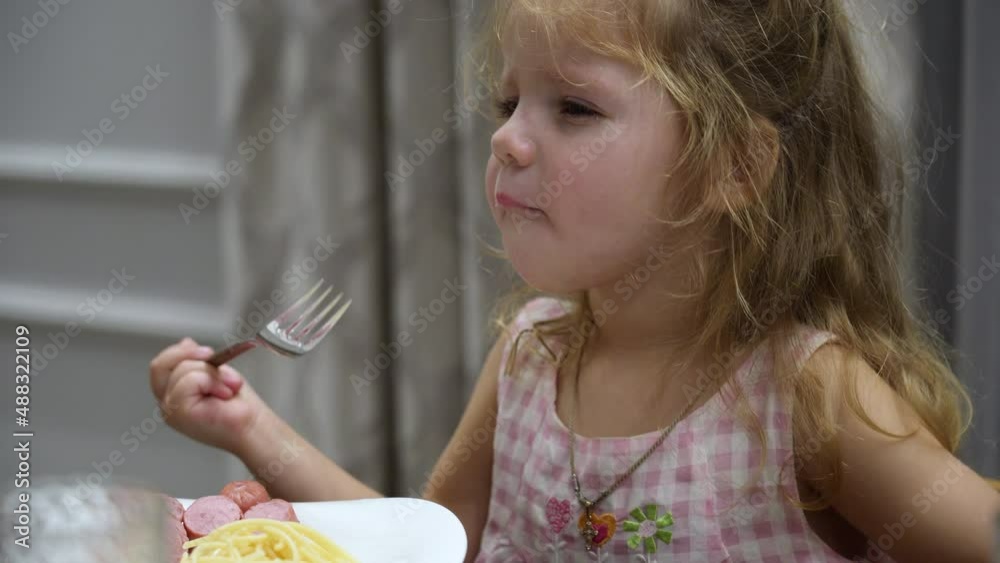 offended, capricious little girl with tears in her eyes sobs heart-rendingly and calls her mother while sitting at the dining table and eating a sausage in the living room