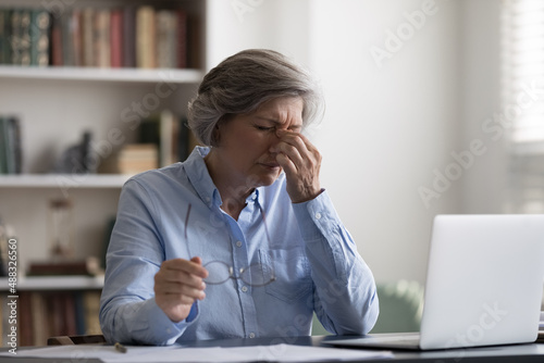Tired middle aged woman taking off eyeglasses, suffering from painful feelings or having blurry eyesight, having astigmatism problem. Stressed due to long computer workday old lady relieving eyes pain photo