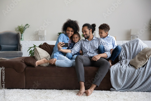 Happy loving young African American family using cellphone, caring parents and little kids son daughter recording funny video or making selfie photo, watching funny content online, tech addiction.