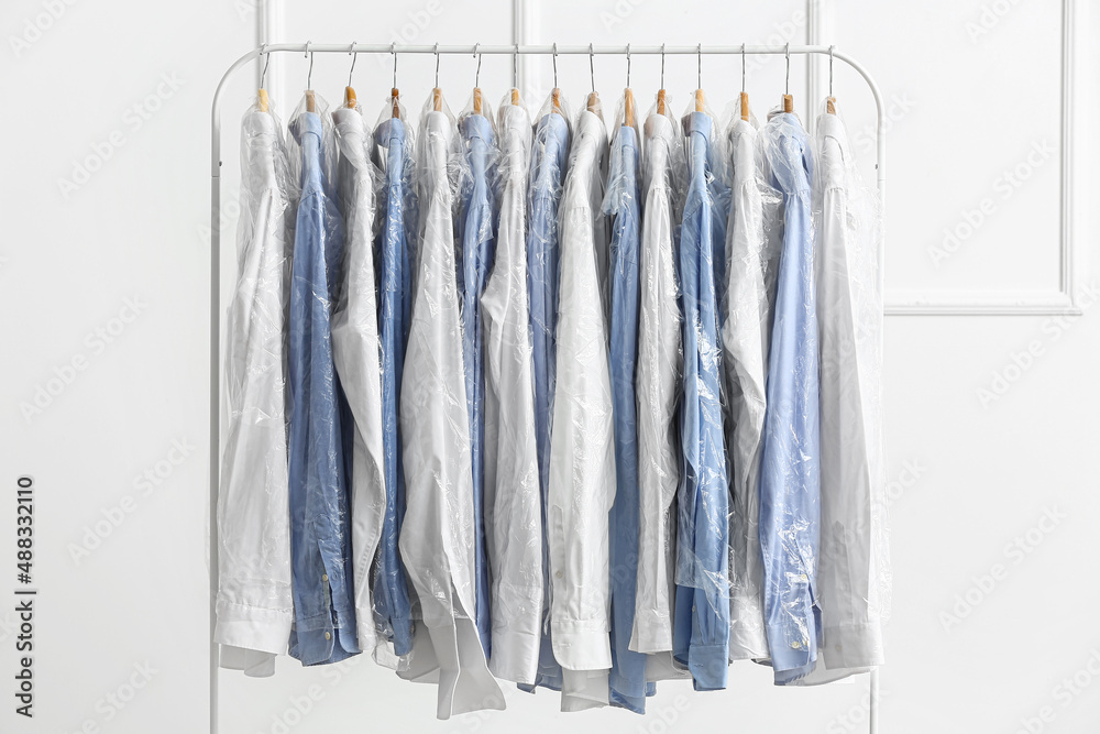 Rack with clean shirts in plastic bags near light wall