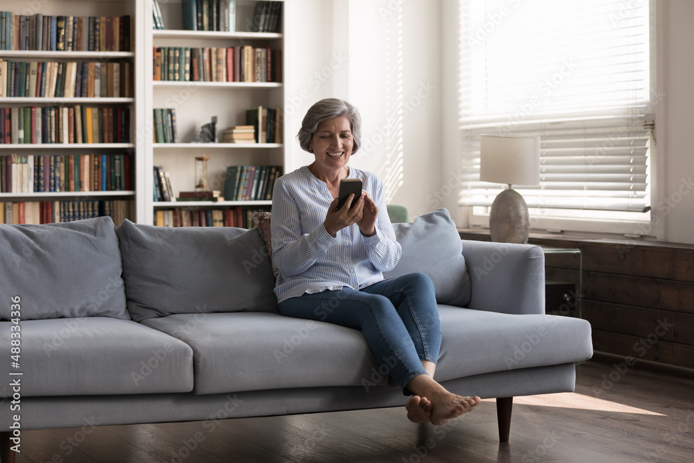 Full length happy middle aged 50s woman using smartphone apps, sitting on comfortable sofa at home, enjoying communicating in social network, playing games. shopping or holding video call conversation